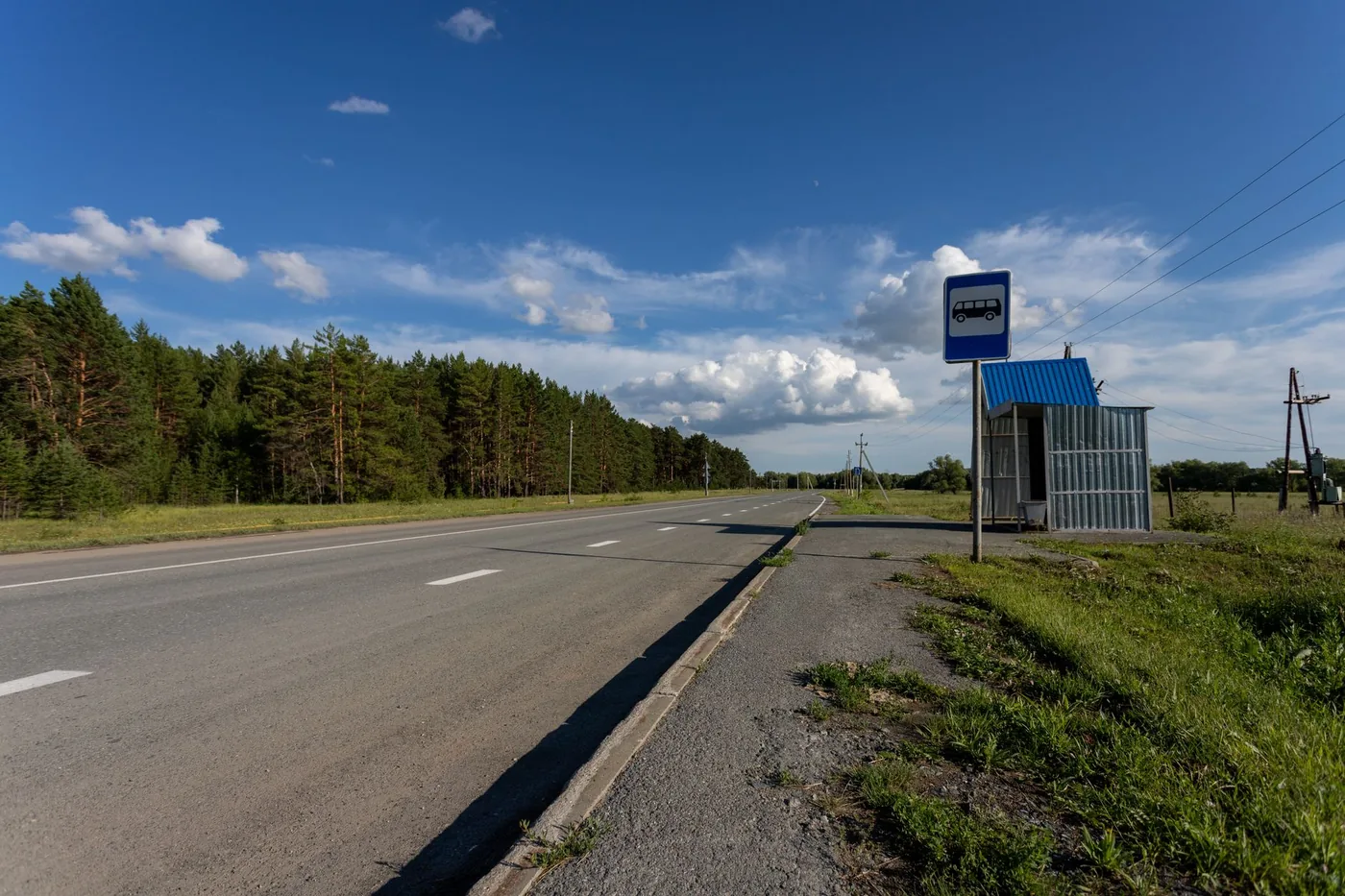 Empty rural road with a bus stop in Sinitsyna, Russia, surrounded by green trees and blue skies.