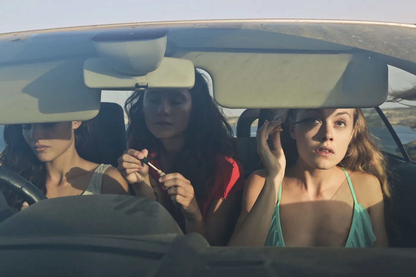 Three young women carpooling and applying makeup inside a convertible car.