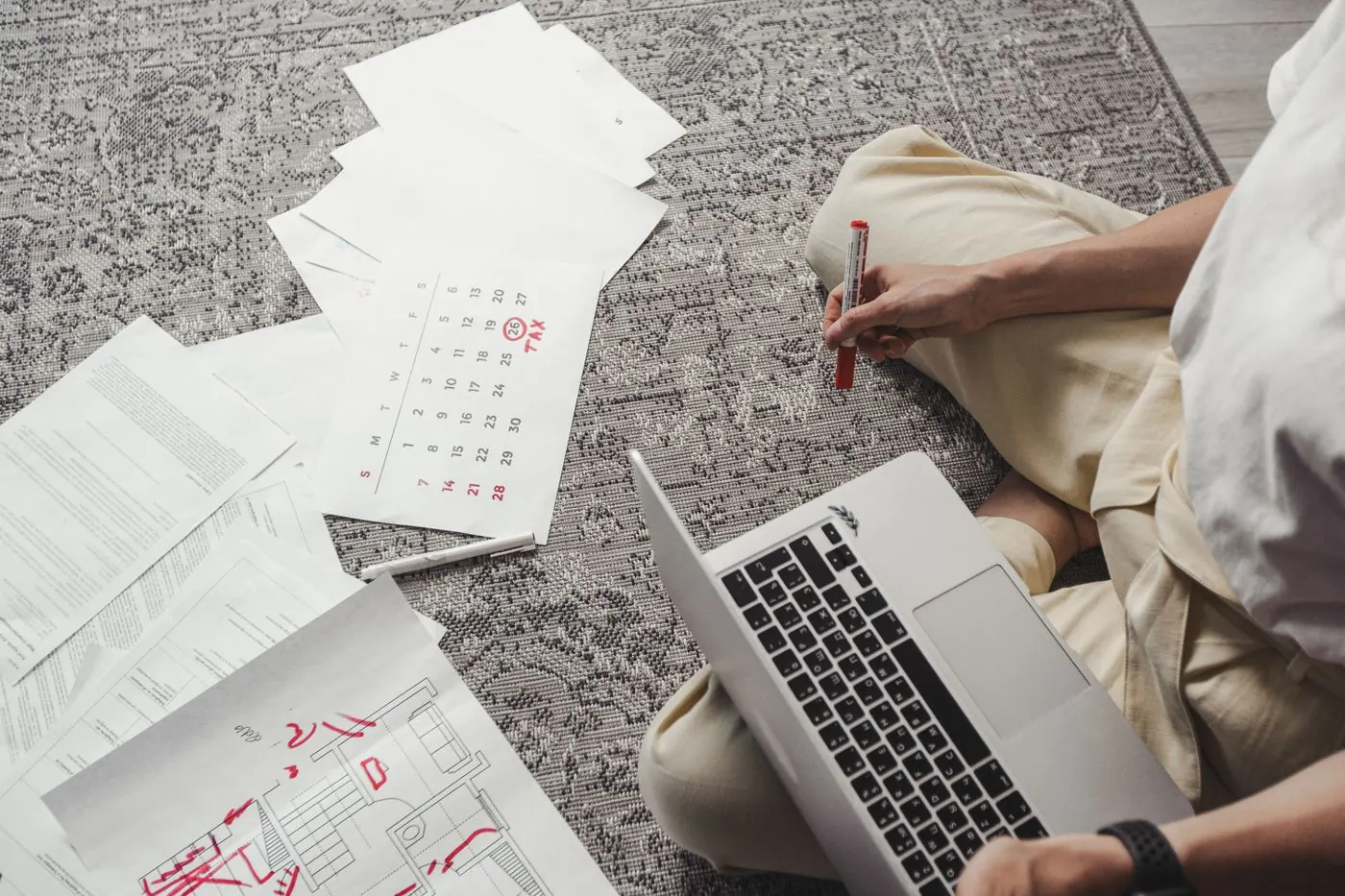 A person with a laptop on a rug, holding a red marker over a printed monthly calendar.
