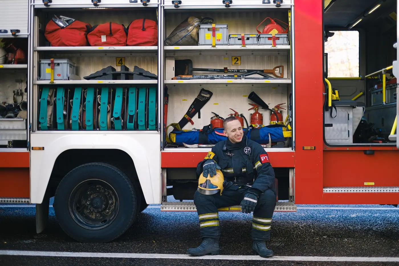Firefighter in uniform sitting on the running board of a fire truck, smiling.