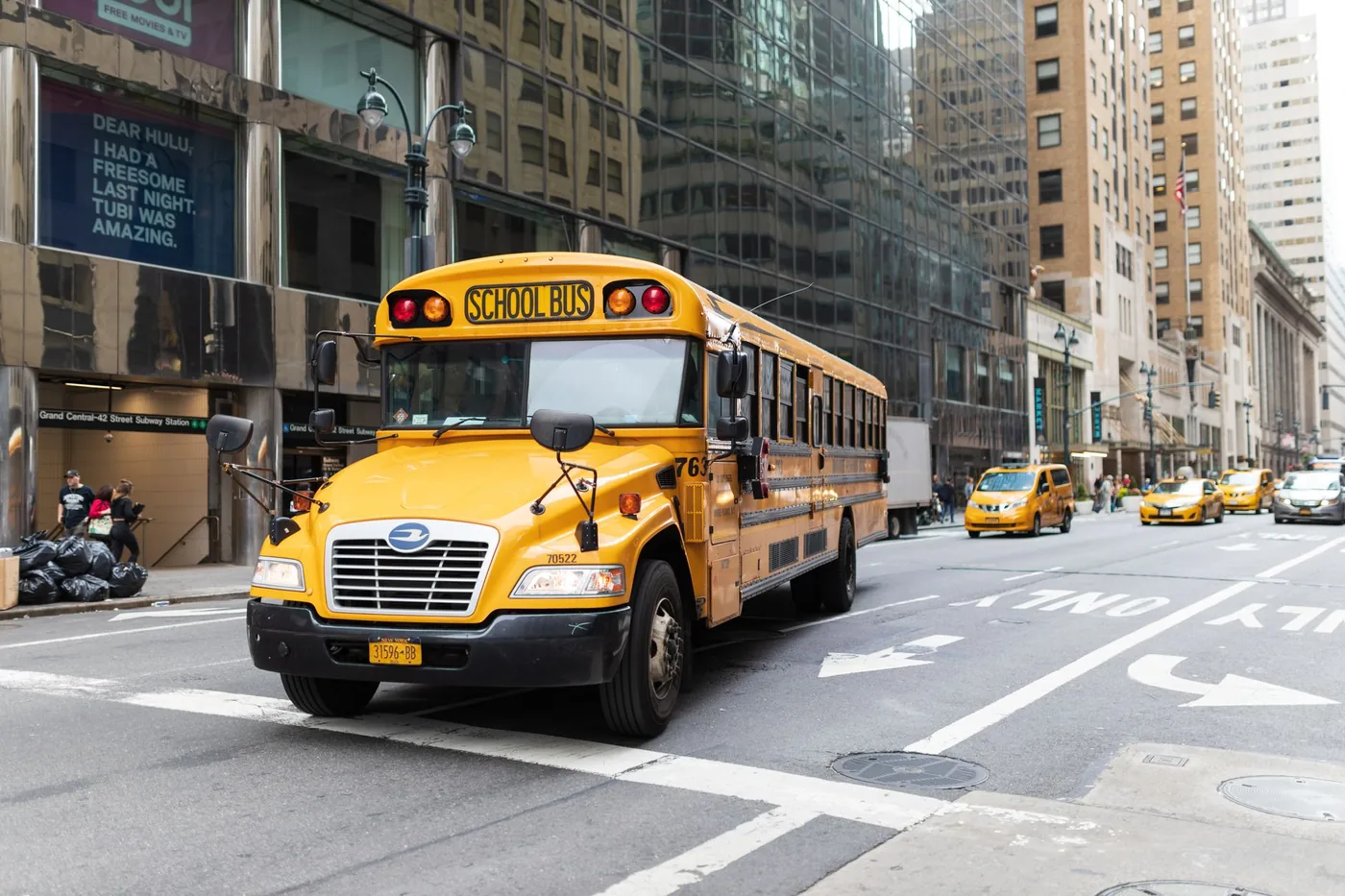 A yellow school bus navigates a busy New York City street amidst tall buildings and taxis.
