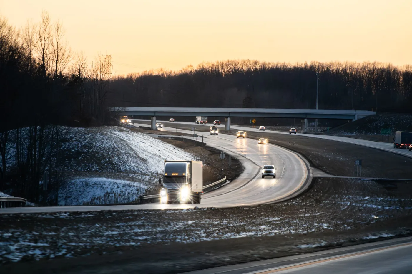 Cars and trucks travel on a highway at dusk with light snow on the ground.