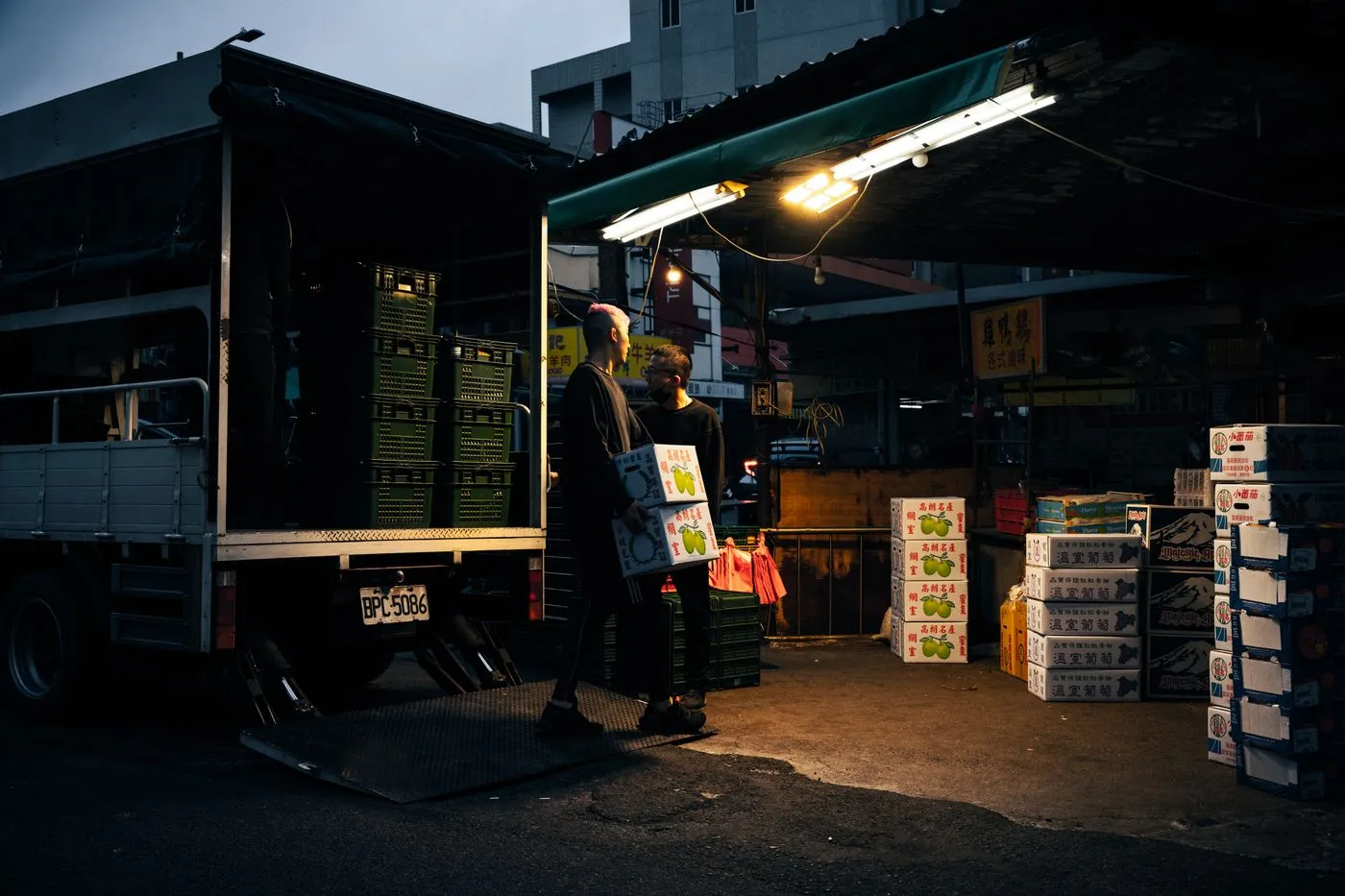 Two workers unloading boxes from a delivery truck under work lights at dusk, as one shift hands over to the next.