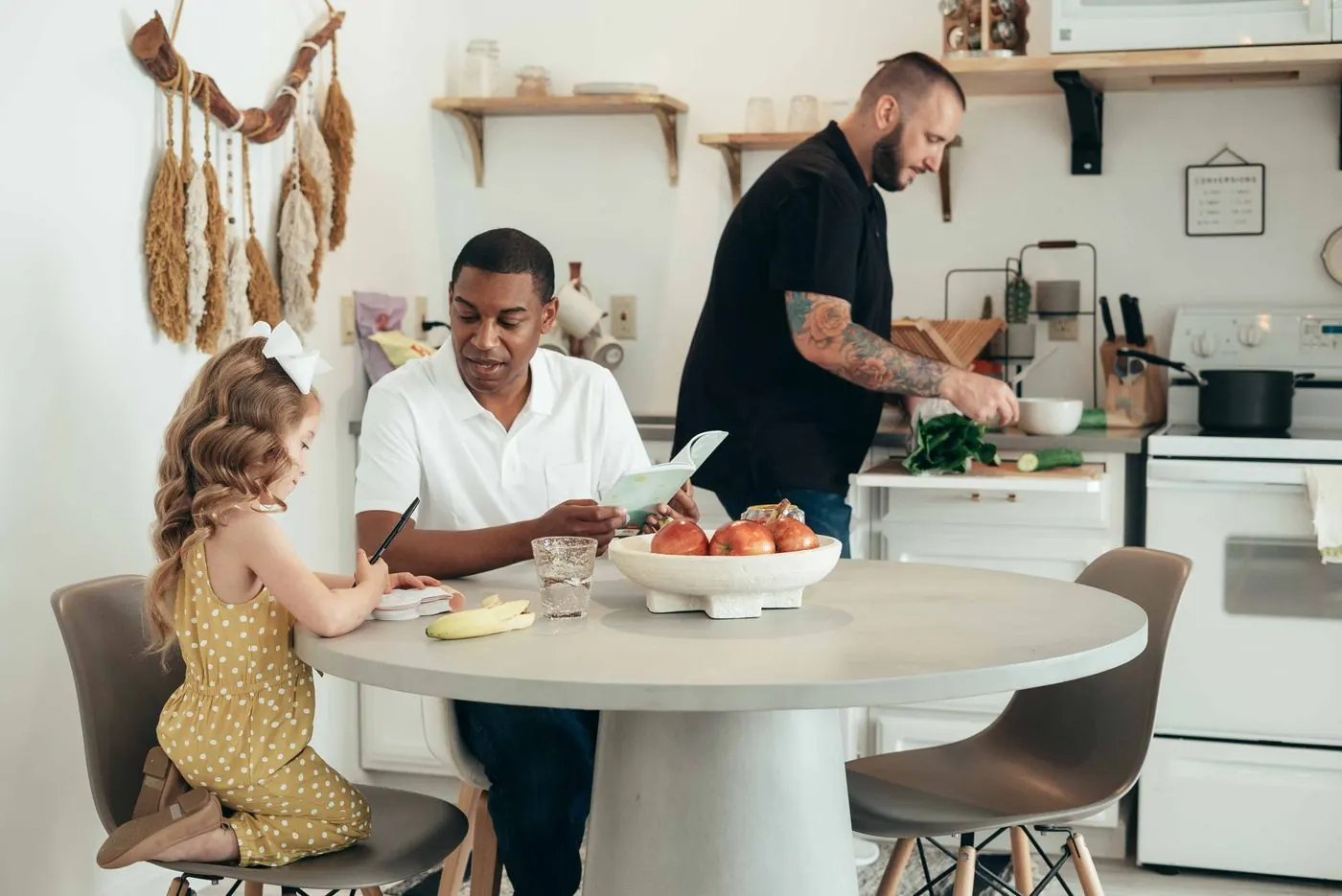 A young girl writing at a kitchen table beside her father while another man prepares food at the counter.